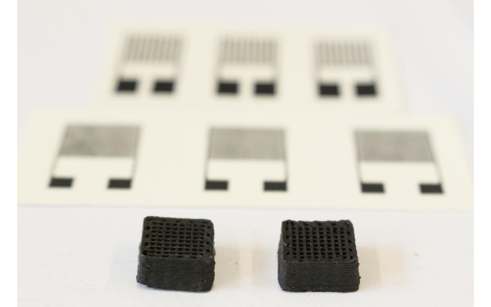 Neuartiger Werkstoff für gedruckte Schaltungen: Zwei Testquader von 1 cm Breite aus dem 3D-Drucker. Im Hintergrund sieht man die gedruckten elektronischen Sensoren.